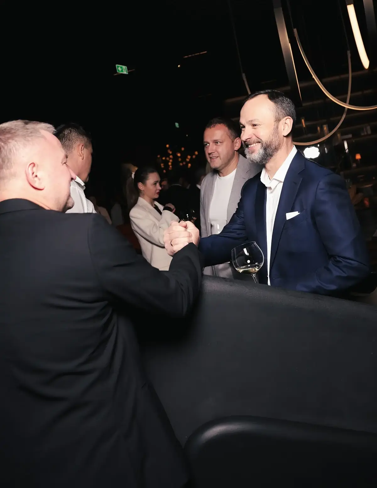 Two people shaking hands, glasses in hand, in a dimly lit room.